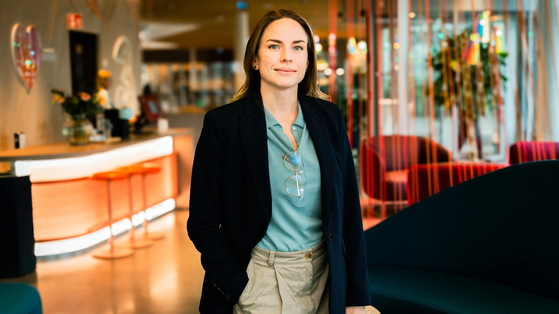 Kvinna i blå kavaj och beige byxor står i ett modernt kontor med färgglada möbler och växter i bakgrunden.