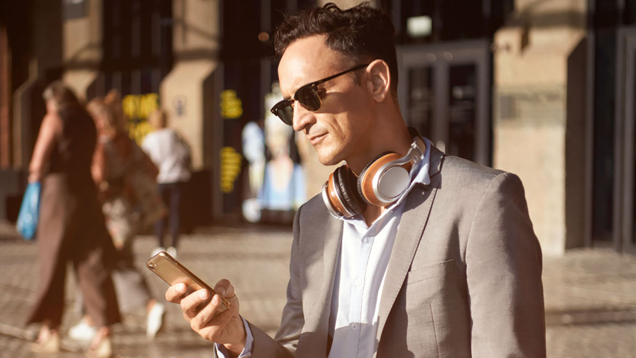 Man i grå kavaj, svarta solglasögon och hörlurar runt halsen hållandes i en mobil i ena handen.