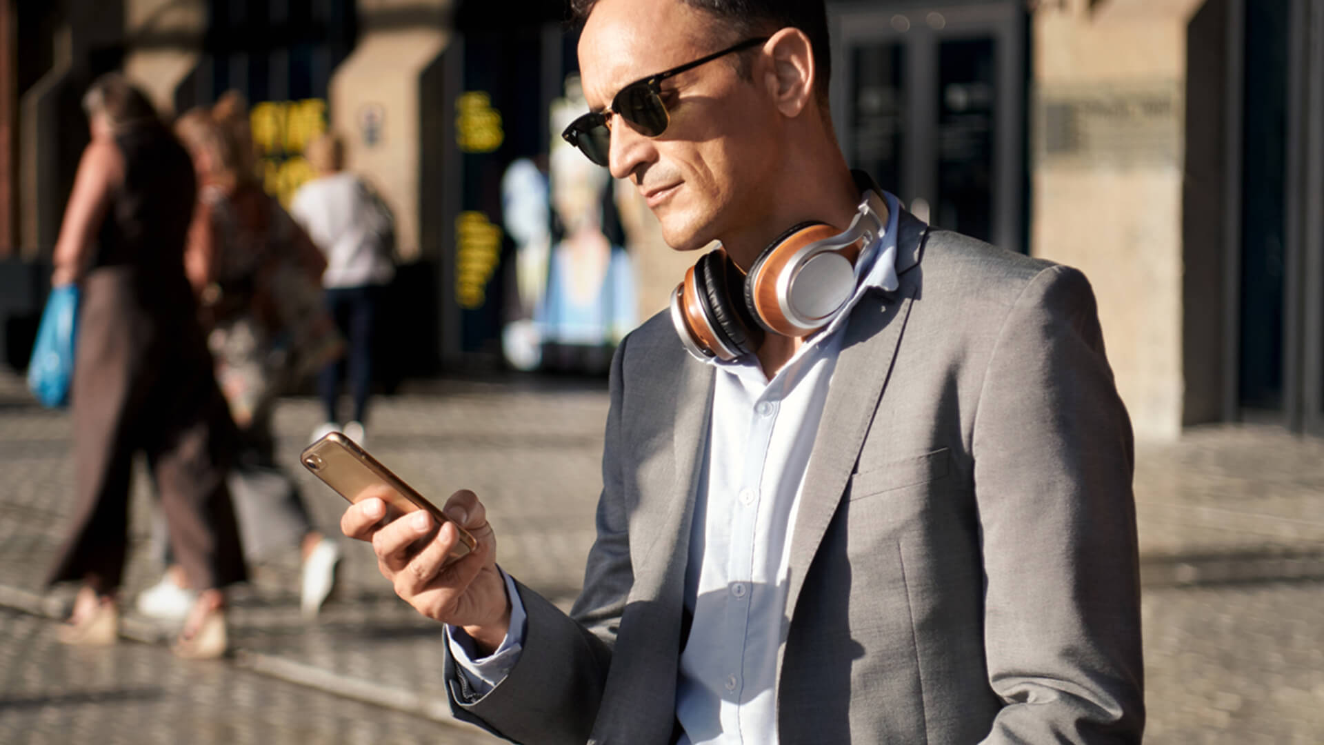 Man i grå kavaj, svarta solglasögon och hörlurar runt halsen hållandes i en mobil i ena handen.