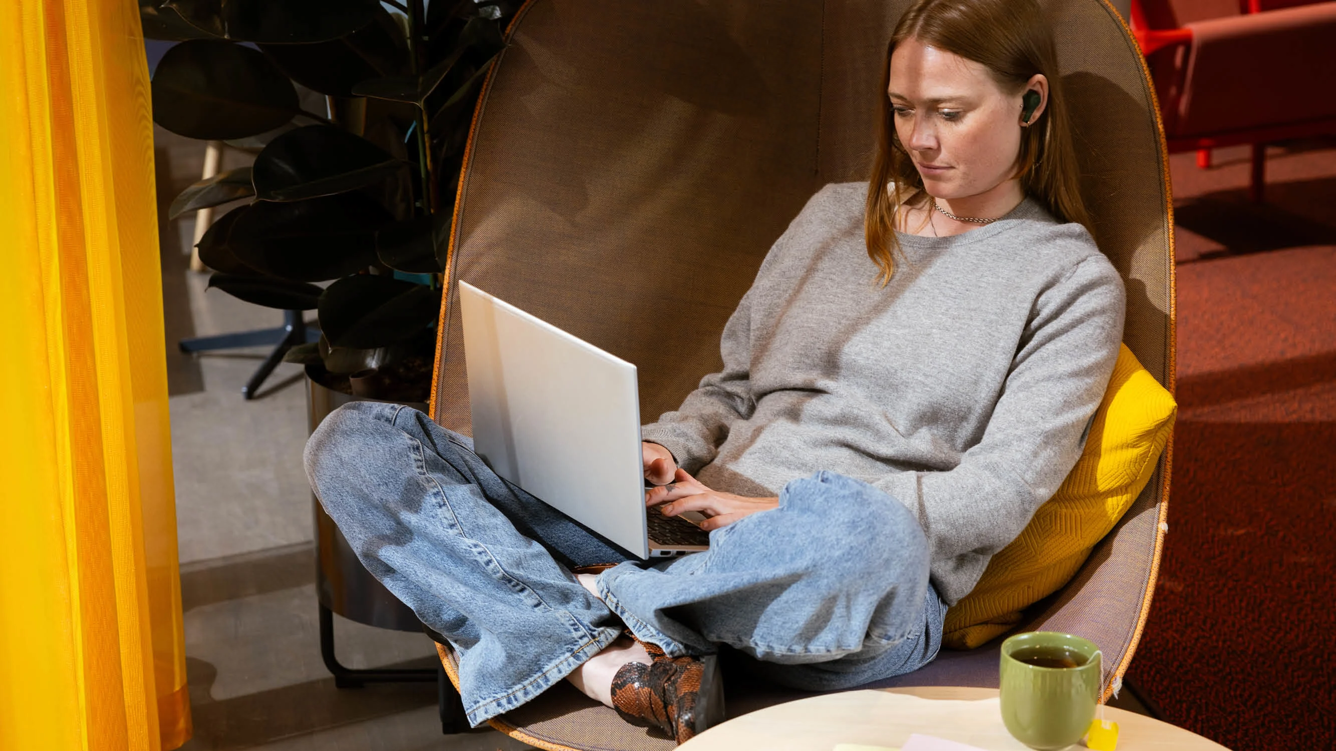 En kvinna i grå sweatshirt och jeans har krupit upp i en fåtölj för att jobba med sin dator