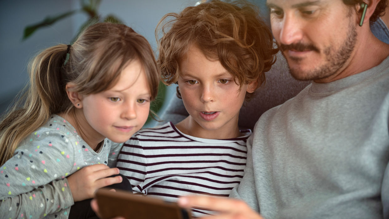 Två barn, en tjej och en kille sitter tillsammans med sin pappa och tittar på en mobiltelefon.