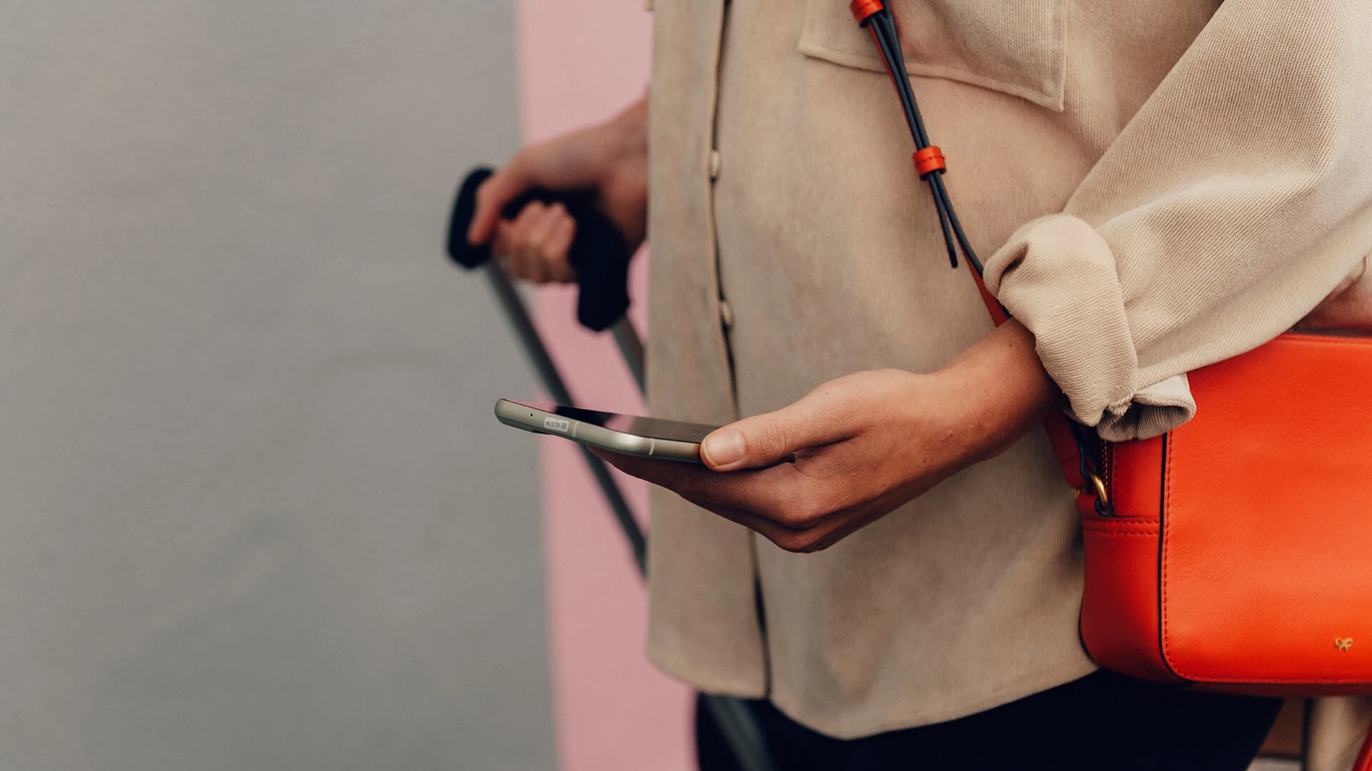 Person i beige skjortjacka håller en mobil i ena handen och drar en resväska i den andra. En orange väska hänger över axeln.