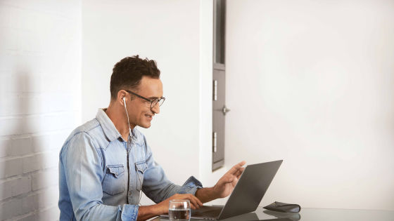 En man i mörkt hår, glasögon och ljusblå jeansskjorta sitter vid ett skrivbord och jobbar på en dator.