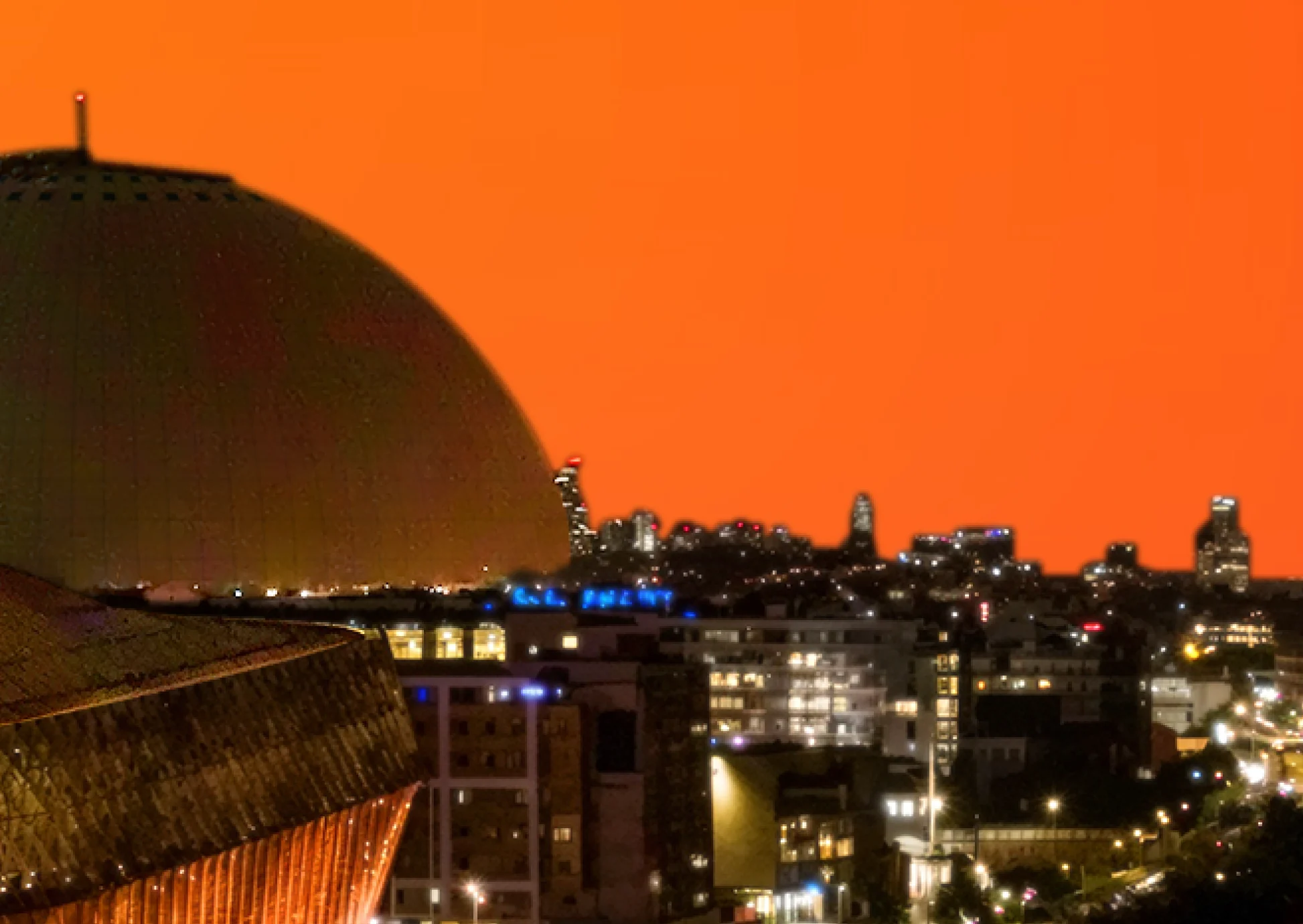 Stockholm skyline med Avicii Arena (Globen) i förgrunden mot en orange kvällshimmel. Stadens ljus lyser upp byggnaderna i bakgrunden