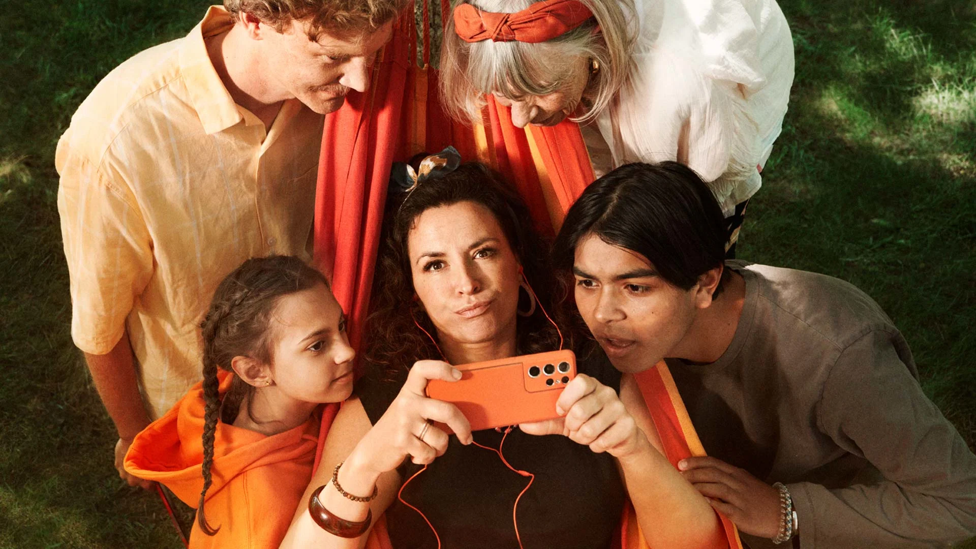 En kvinna ligger i en orange hängmatta med en mobil, personer står unt omkring fr att också titta.