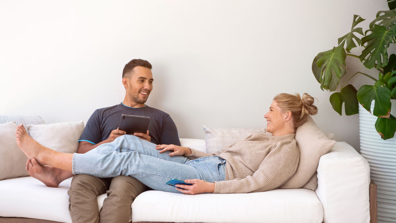 En man i mörkt hår sitter i en soffa med en surfplatta i händerna. En kvinna i blont hår ligger i soffan med benen över honom. Hon håller i en mobiltelefon. 