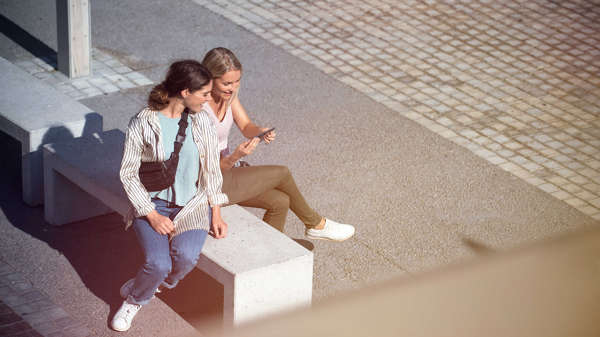 Två kvinnor sitter tillsammans på en grå betongbänk och samtalar. Den ena kvinnan visar den andra något på sin mobil.