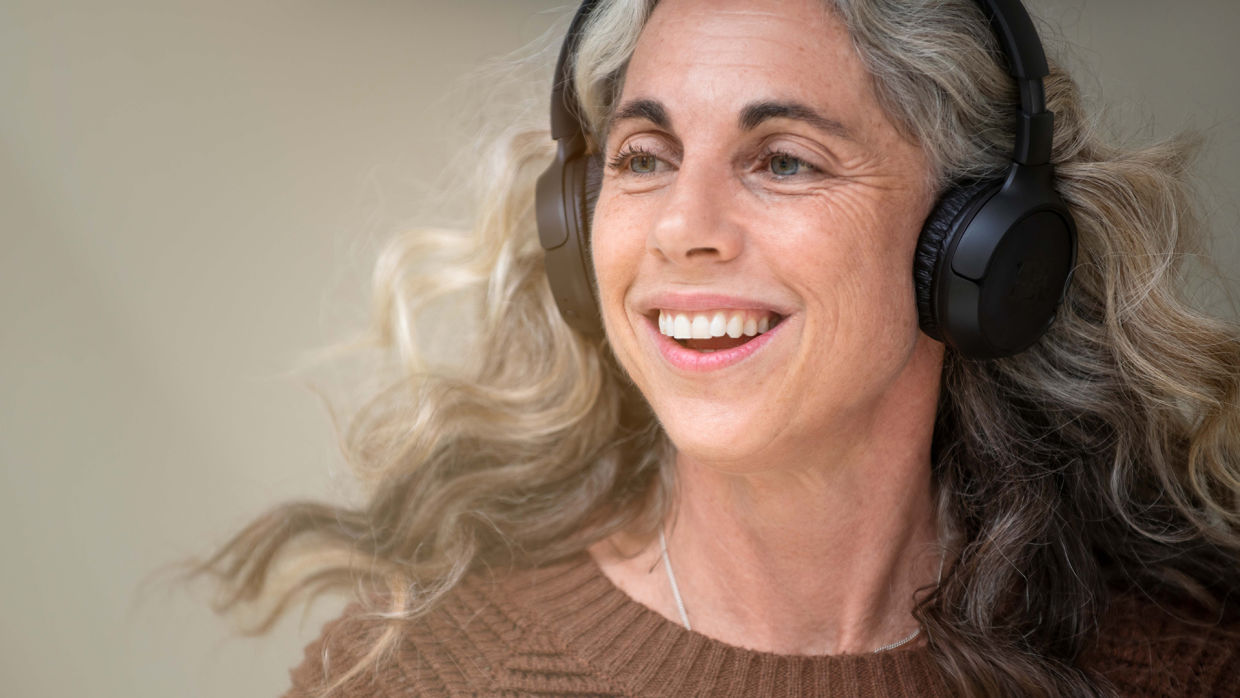 Leende kvinna med grått, yvigt hår lyssnar på musik i ett par on ear-hörlurar.