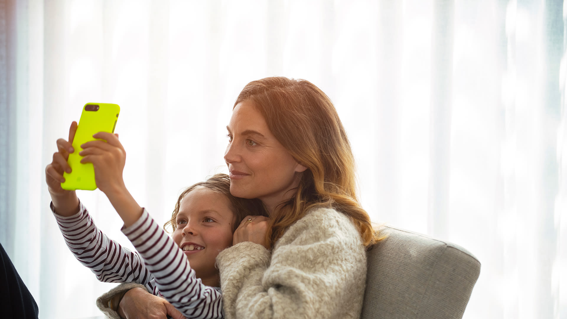 Kvinna i rödbrunt hår sitter i en soffa. I knät sitter ett barn och håller upp en mobil. Båda tittar på skärmen.
