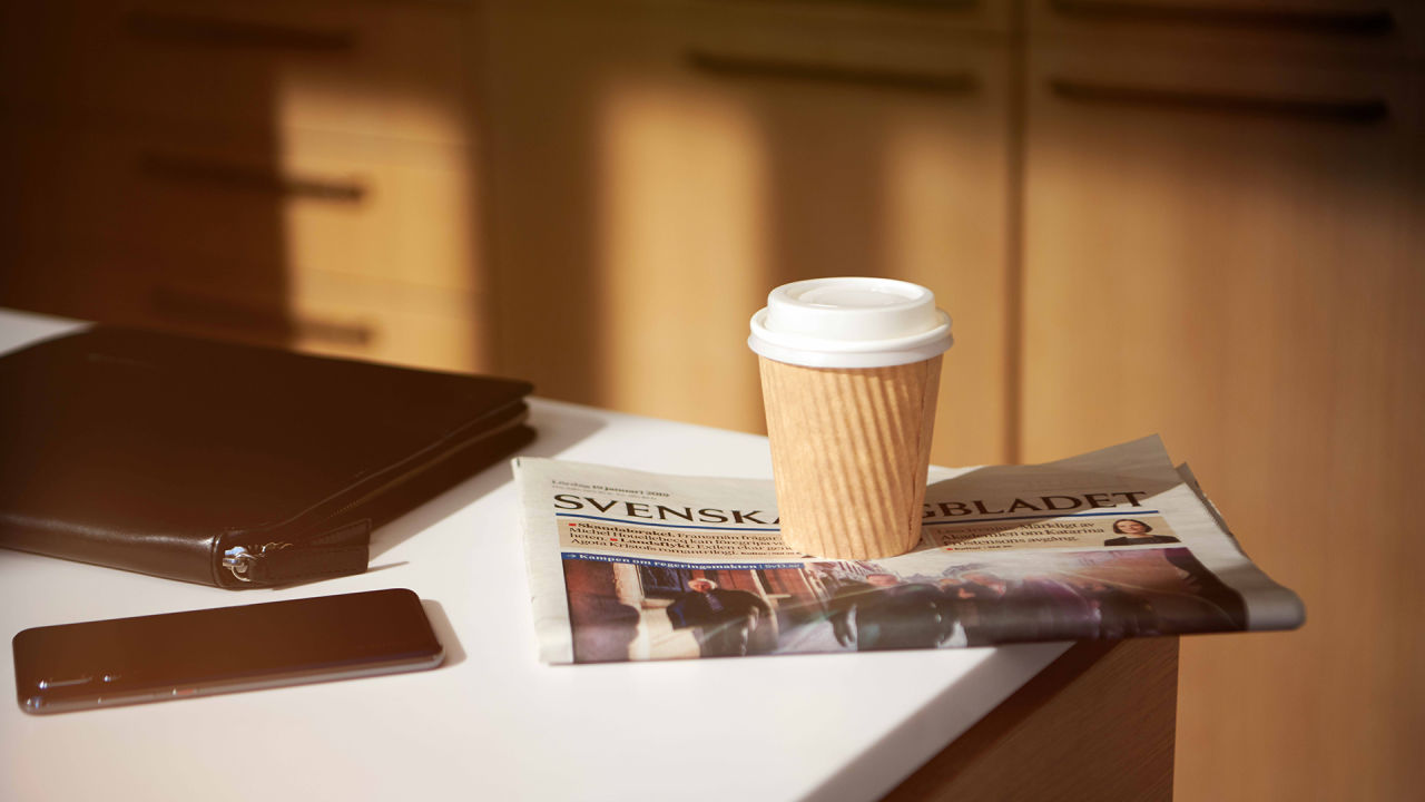 En vit bänkskiva där det ligger en mobiltelefon, en surfplatta i ett svart fodral, een tidning och en kopp kaffe i take away-mugg.