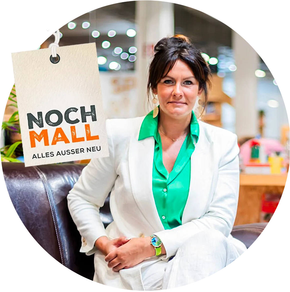 Eine Frau in einem weißen Anzug und einer grünen Bluse sitzt auf einem braunen Ledersofa in einem modernen Geschäft. Neben ihr steht ein Schild mit der Aufschrift NOCH MALL ALLES AUSSER NEU. Im Hintergrund sind Regale und Waren zu sehen.