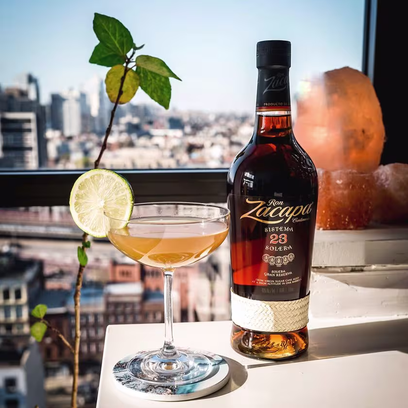 La imagen muestra una copa rellena con el cóctel Daiquiri y una botella de ron Zacapa 23. De fondo, una ventana amplia donde se puede observar la ciudad.