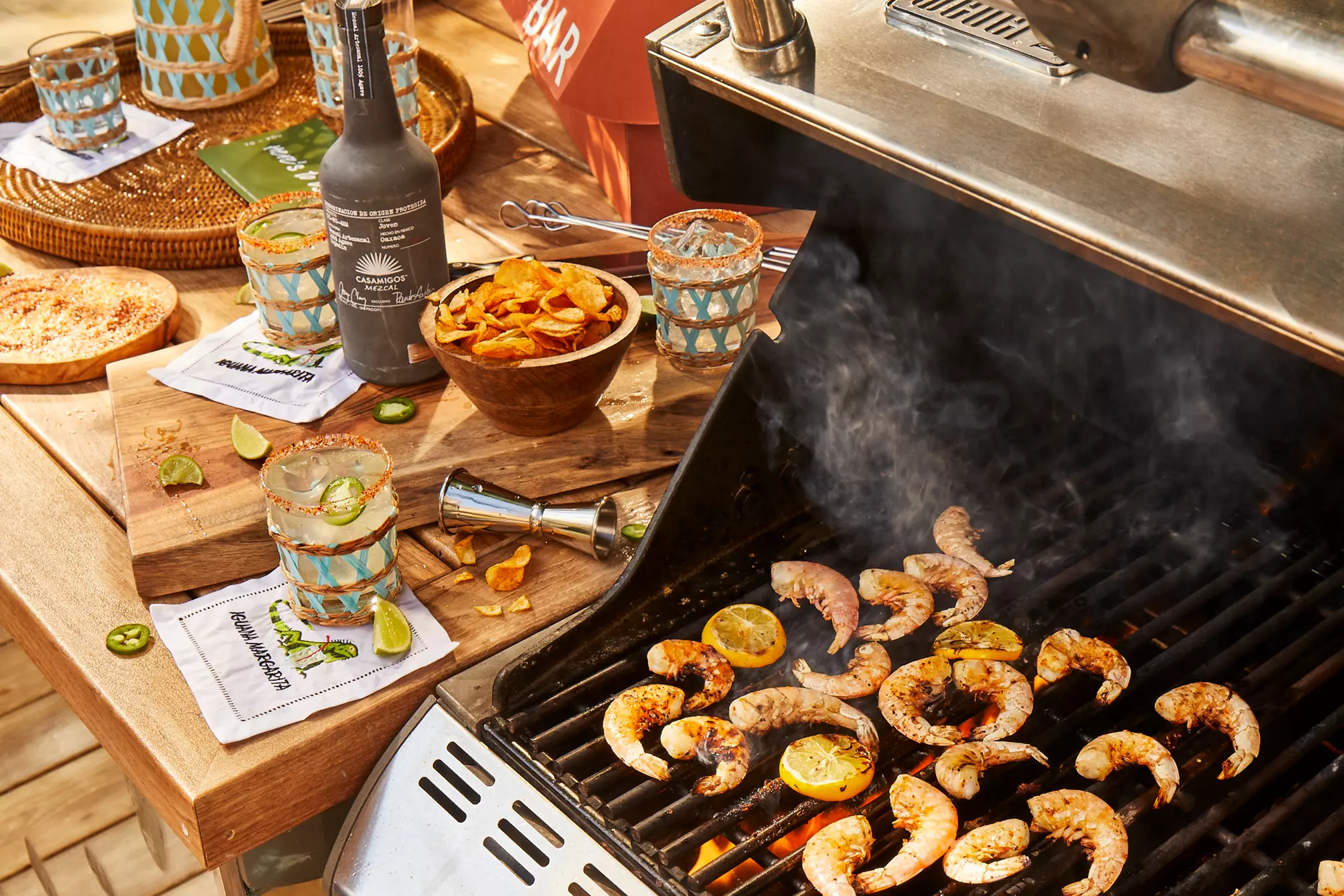 Shrimp and lime slices grilling on a barbecue with drinks, chips, and napkins arranged on a nearby wooden table.