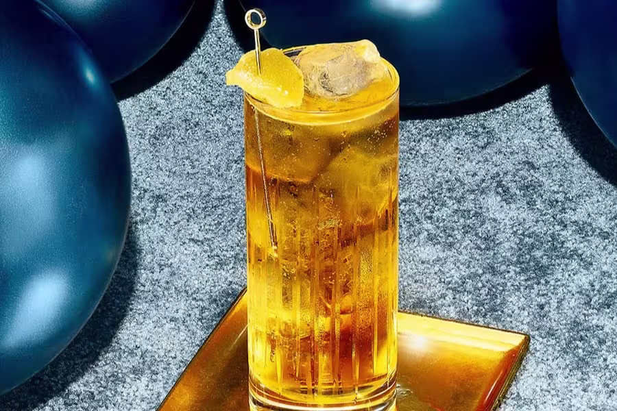 A tall glass of amber-colored cocktail with ice and a lemon garnish sits on a gold tray, with blue balloons in the background.
