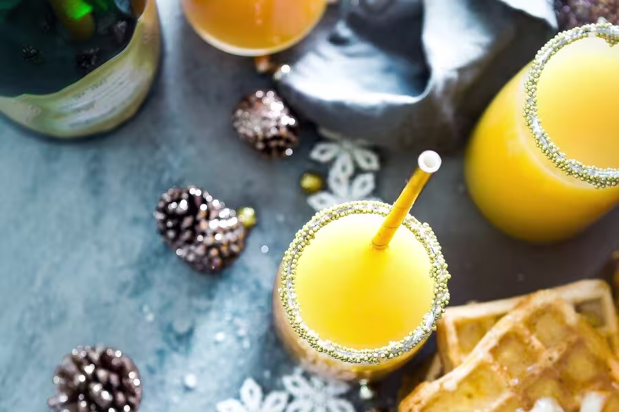 A flat lay of orange drinks with gold sugar rims, a straw, waffles, pine cones, and a bottle on a blue surface.