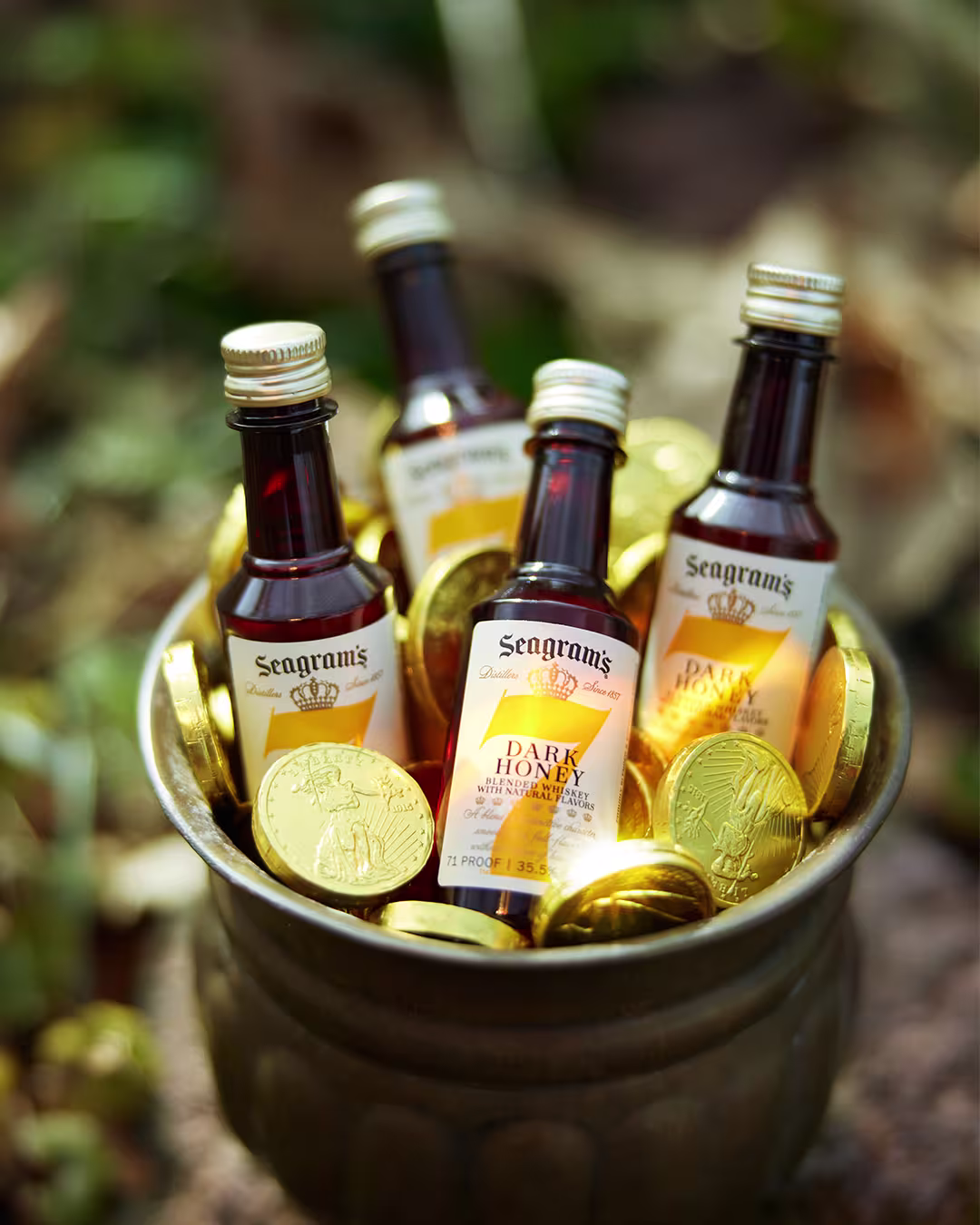 A metal cup filled with Seagram’s Dark Honey mini bottles and chocolate gold coins, set outdoors on grass.
