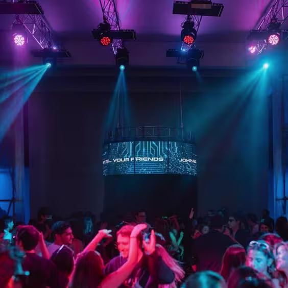 Imagen de discoteca, gente bailando , luces azules y violetas. De fondo la cabina del DJ a altura