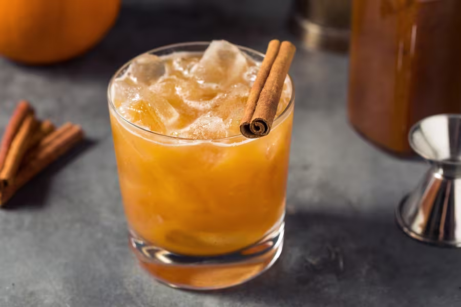 A glass of orange-brown iced cocktail garnished with a cinnamon stick sits on a gray surface, with extra cinnamon sticks and a jigger nearby.