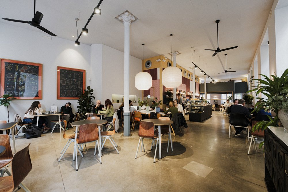 Interior amplio de Federal Café con mesas de madera, columnas blancas, lámparas colgantes, plantas decorativas y ambiente relajado con clientes.