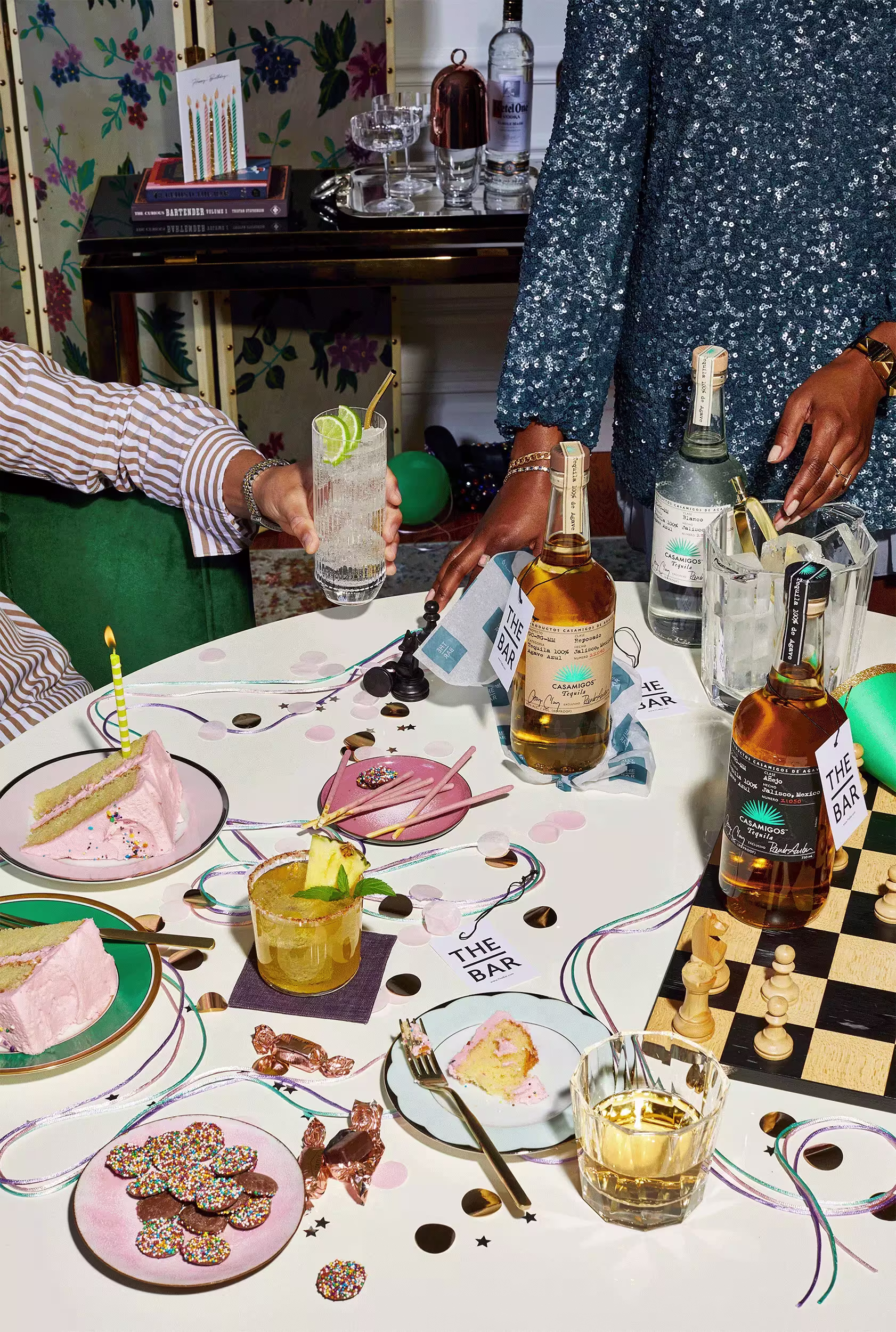 A table set for a celebration with drinks, cake, confetti, party favors, and a chessboard. Two people are standing nearby, one holding a cocktail. Bottles of alcohol are on the table.
