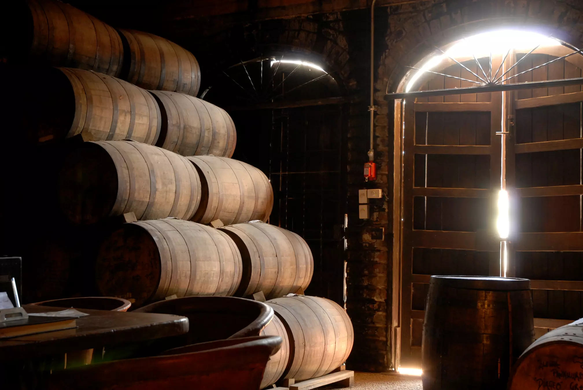 Stacks of wooden barrels inside a dimly lit cellar with sunlight streaming through a partially open wooden door.