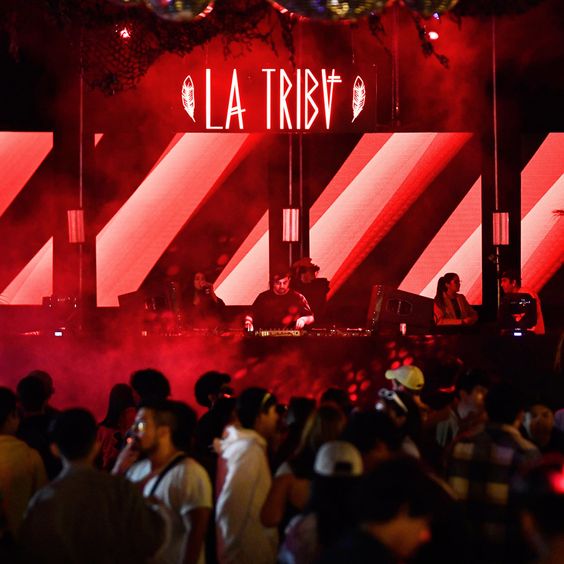 Imagen de la cabina del DJ con cartel de neón rojo que lleva el título de la fiesta: "La Tribu", por delante se ve a la gente bailando entretenida.