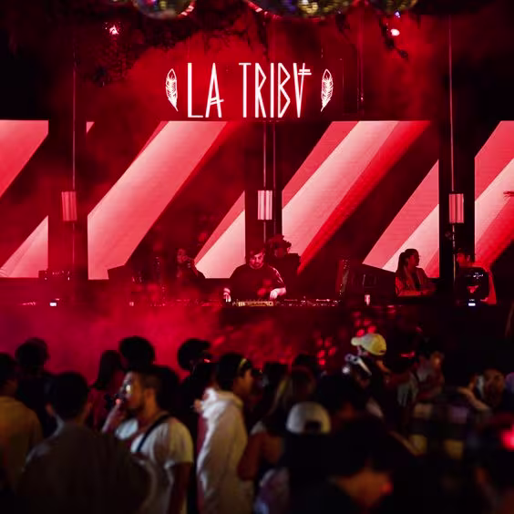 Imagen de la cabina del DJ con cartel de neón rojo que lleva el título de la fiesta: "La Tribu", por delante se ve a la gente bailando entretenida.