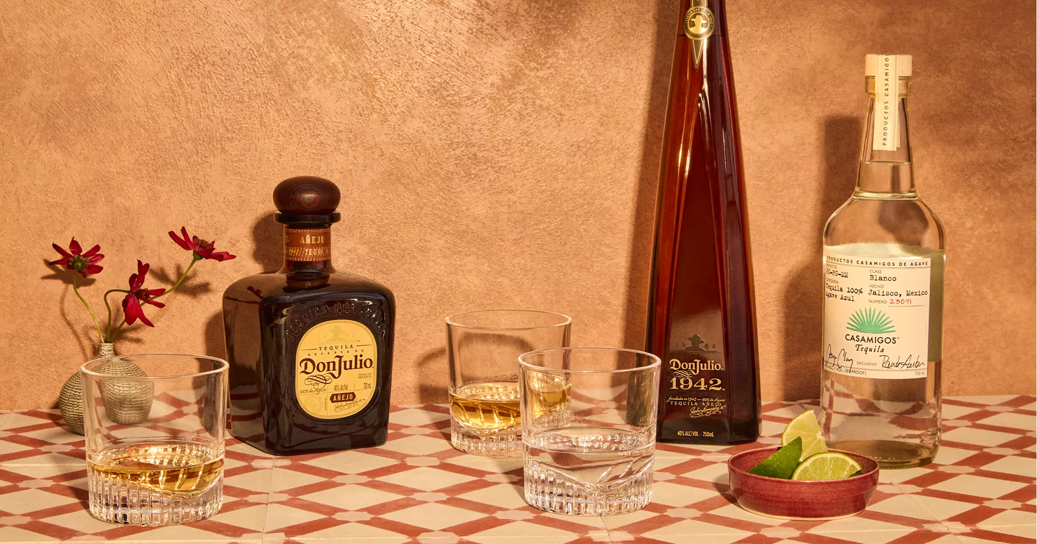 Three bottles of tequila with labels visible, three glasses with tequila, a small vase with red flowers, and lime wedges on a checkered surface against a tan background.