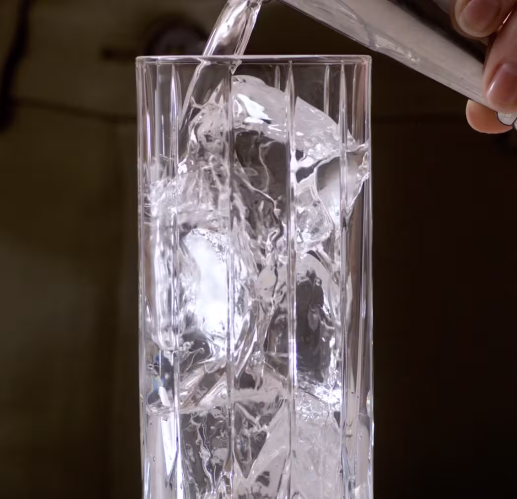A tall glass filled with ice cubes, as a clear liquid is being poured into it from a small container, illustrating a step in preparing a cocktail.
