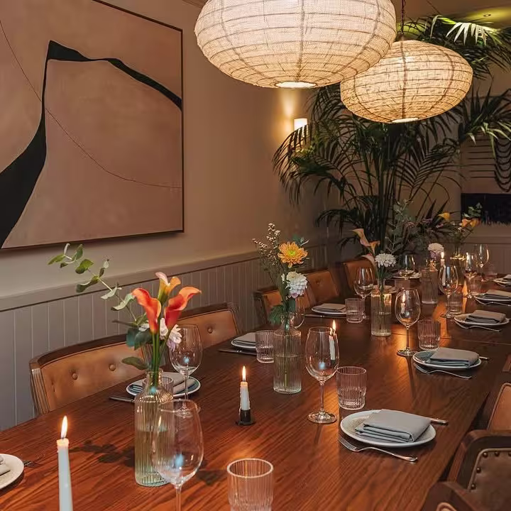 La imagen muestra una elegante mesa larga decorada con flores frescas en jarrones de vidrio, rodeada de sillas de madera, en una habitación iluminada con lámparas colgantes de estilo moderno.