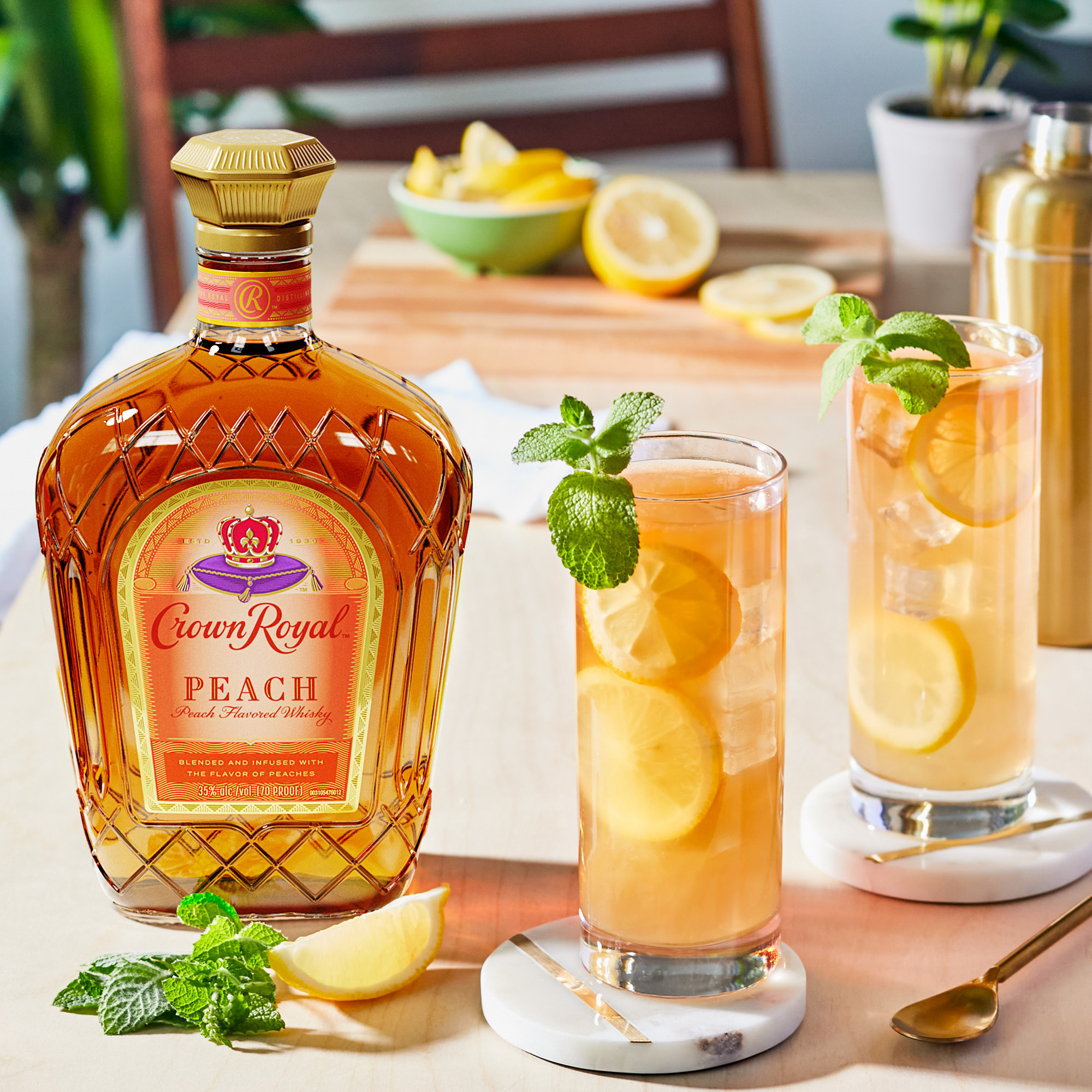 A bottle of Crown Royal Peach whisky sits beside two iced cocktails garnished with lemon slices and mint, with lemon wedges and cocktail-making items in the background.
