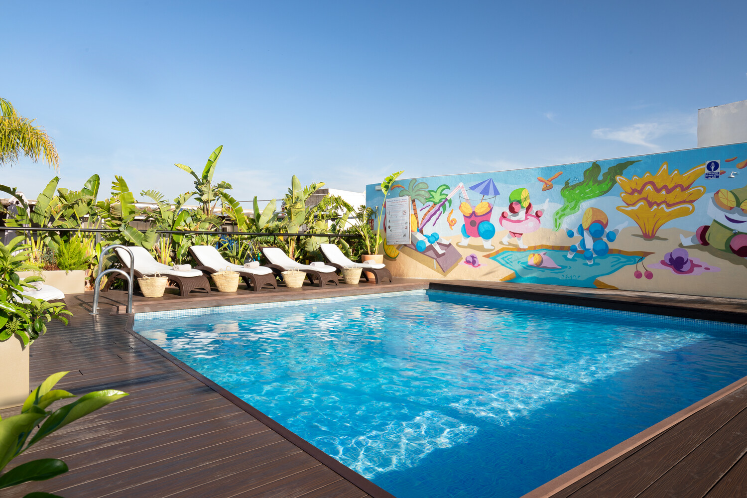 La imagen muestra una zona de piscina al aire libre del Pura vida Sevilla. La piscina está rodeada de varias tumbonas blancas en un lado y un mural colorido en el otro. El mural presenta personajes y diseños vibrantes, estilo caricatura. 