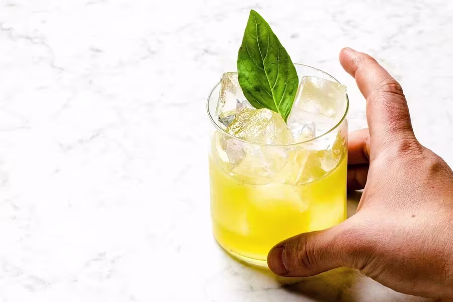A hand holds a glass of yellow iced beverage garnished with a green leaf, on a white marble surface.
