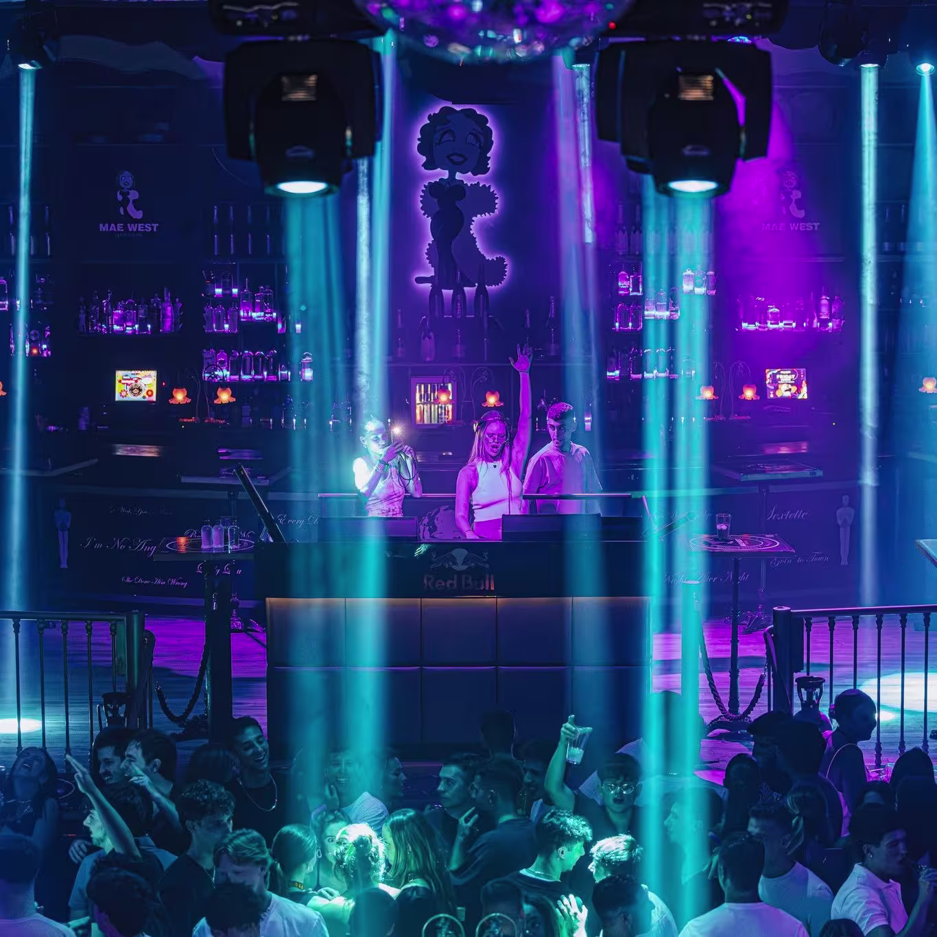 Multitud disfrutando en Mae West, con DJ en el centro iluminado por haces de luz azul y morada. Ambiente festivo con detalles de neón y botellas decorativas en las paredes del club.