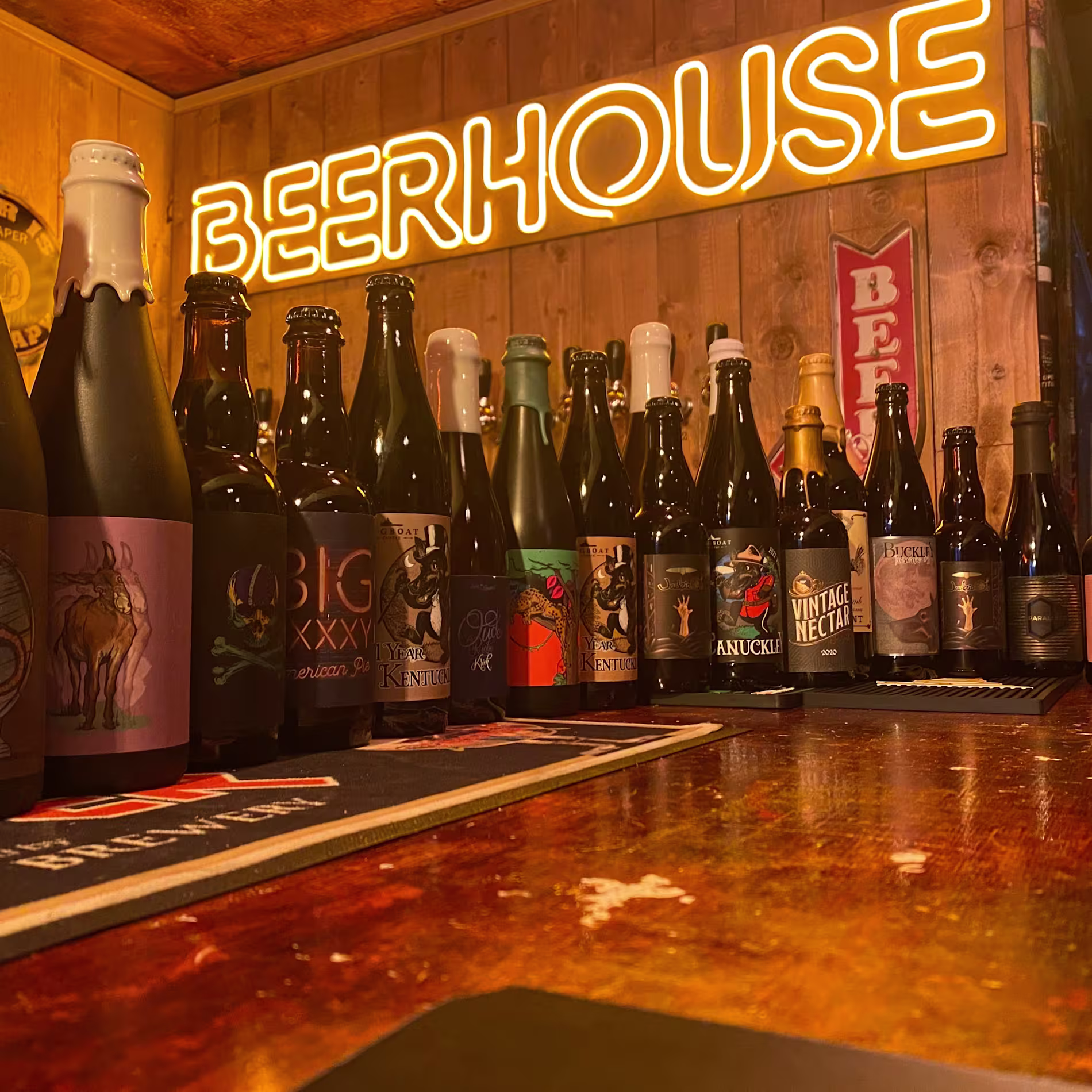 La imagen muestra una colección de varias botellas de cerveza sobre una superficie de madera. En el fondo, hay un letrero de neón que dice “BEERHOUSE”. Un bar especializado en cervezas artesanales.