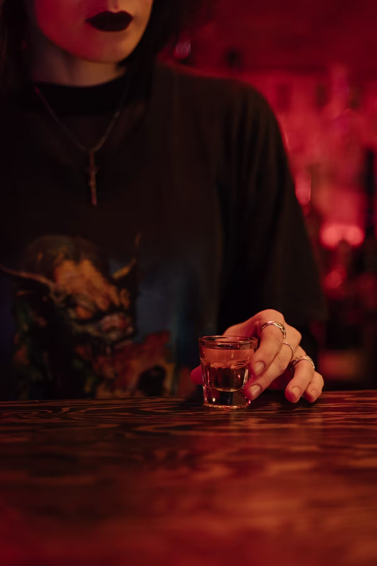 A person with dark nail polish and rings holds a shot glass on a wooden bar counter, wearing a dark shirt and a cross necklace in a dimly lit setting.