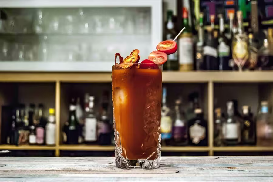 A tall glass of Bloody Mary garnished with cherry tomatoes and a slice of pepper sits on a bar counter with liquor bottles in the background.