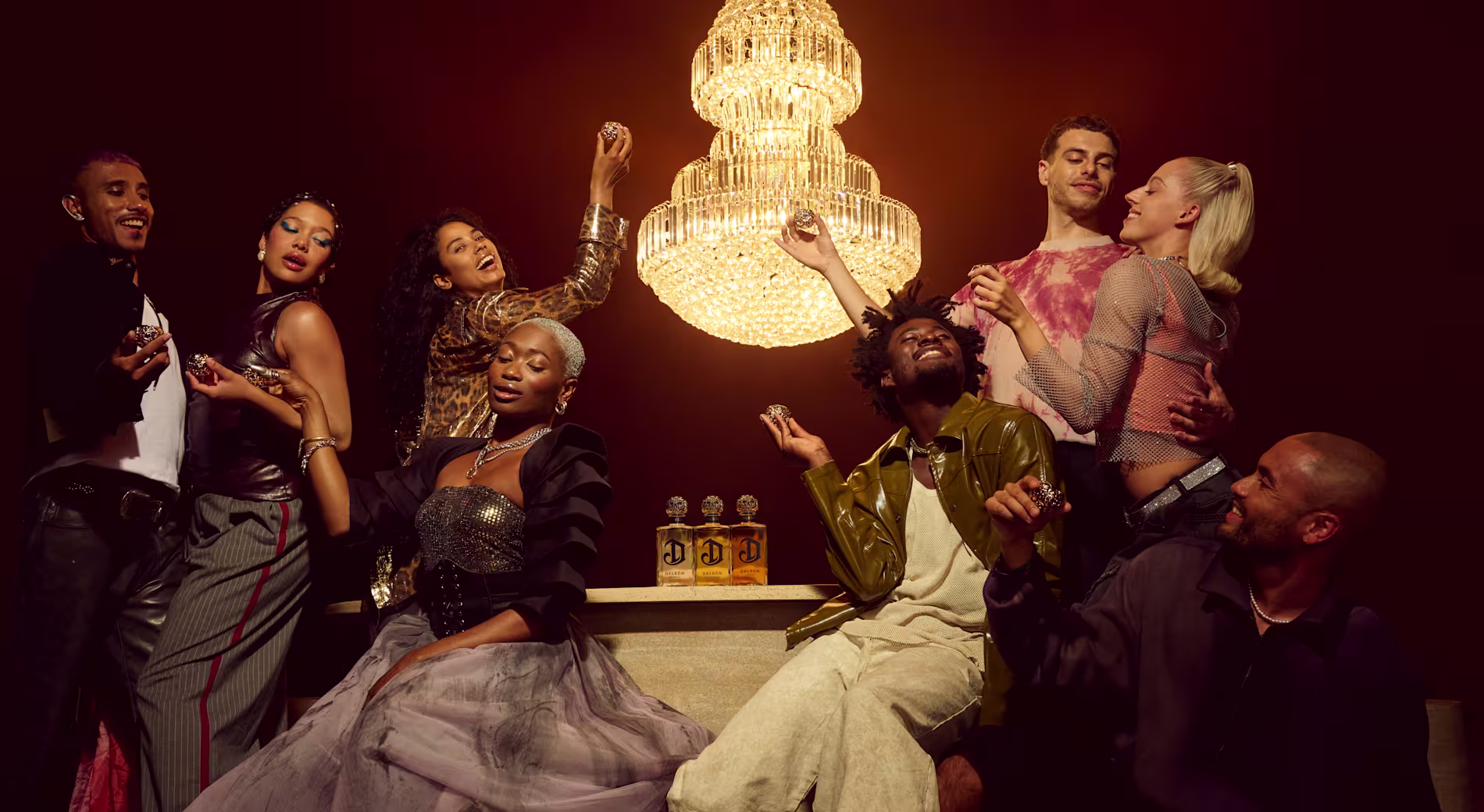 A group of seven stylishly dressed people pose together under a large chandelier, holding drinks and gesturing playfully around a table with three perfume bottles.