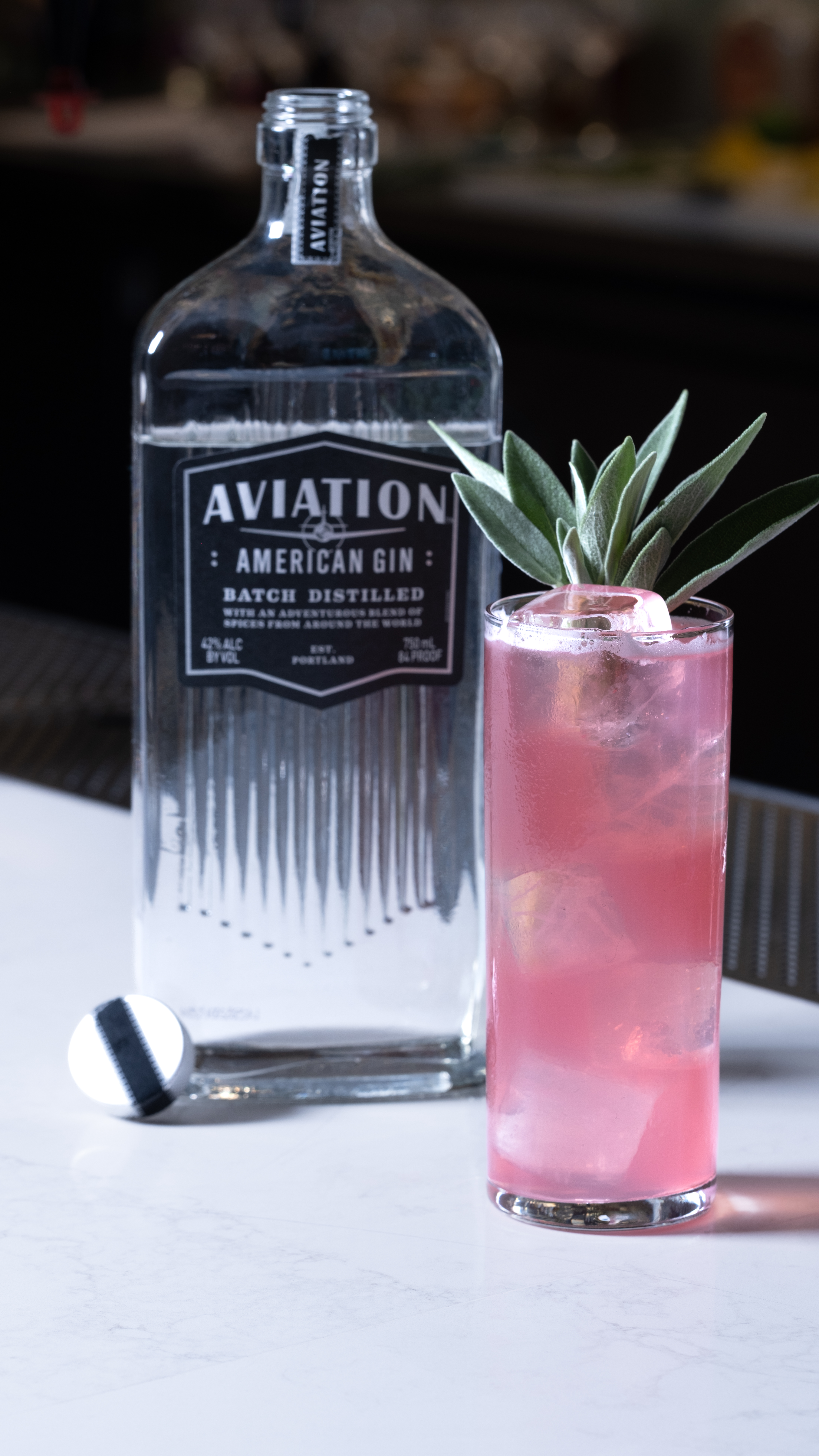A purple coloured cocktail in a highball glass, served next to an Aviation gin bottle