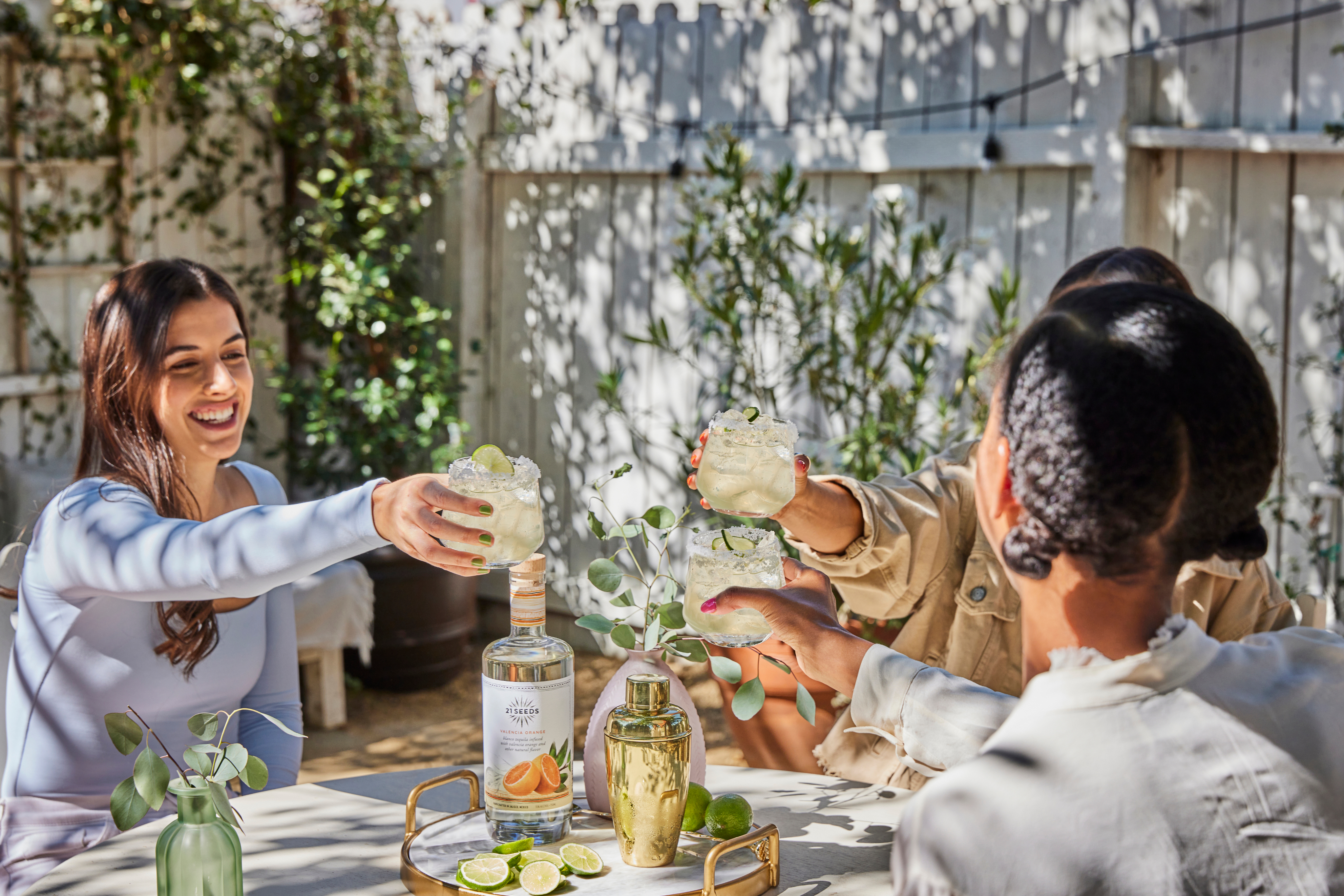 Three people sit around an outdoor table clinking cocktails, with a bottle of tequila, limes, and a cocktail shaker in front of them.
