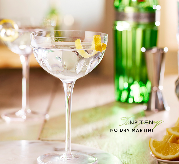 Imagen de trago, en copa alta, decorado con una linea de cascara de limón. Botella de Gin por detas de manera borrosa.
