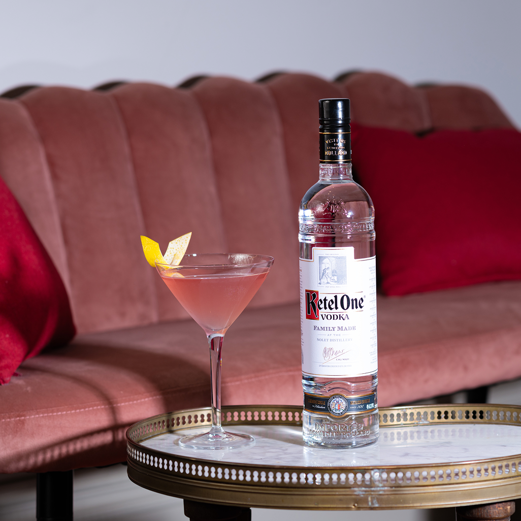 O sticlă de vodcă Ketel One și un cocktail roz cu o garnitură de lămâie pe o masă rotundă în fața unei canapele roz