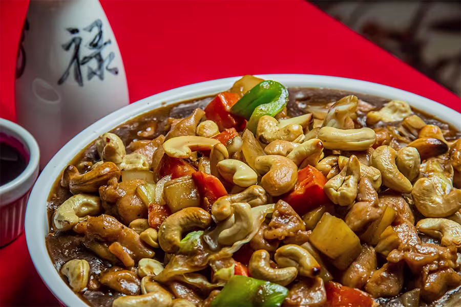 A bowl of stir-fried chicken with vegetables, pineapple, and cashew nuts in sauce, placed on a red table with a small bottle and a dipping sauce cup nearby.