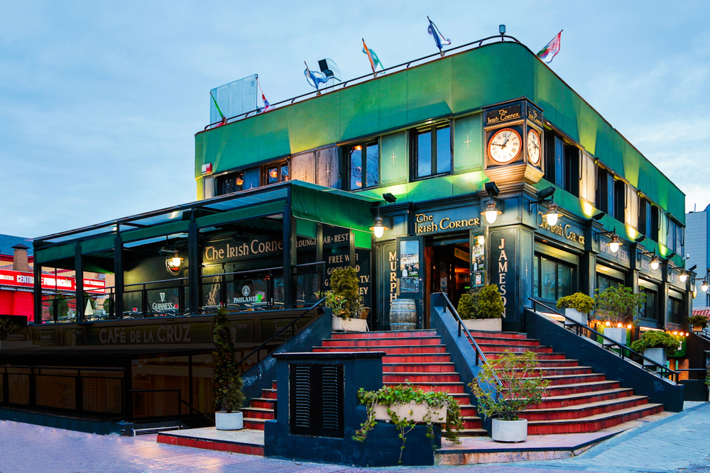 La foto muestra un edificio de dos pisos con una fachada verde, que alberga el pub “The Irish Corner”. Hay varias banderas en el techo, asientos al aire libre en ambos niveles y un reloj cerca del borde del techo. 