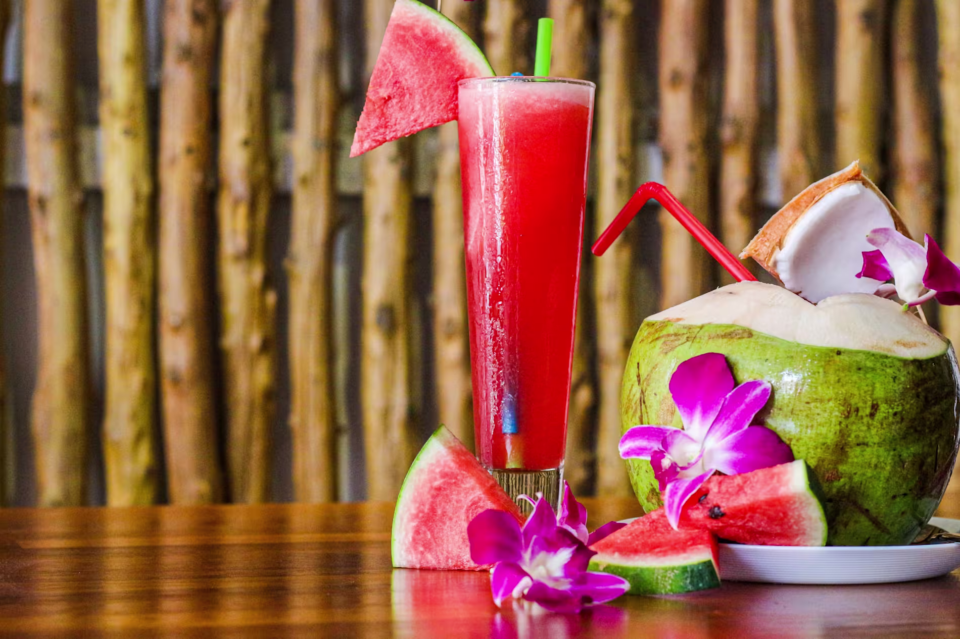 A tall glass of watermelon juice with a watermelon slice, a coconut with a straw and flowers, and watermelon wedges on a wooden table.