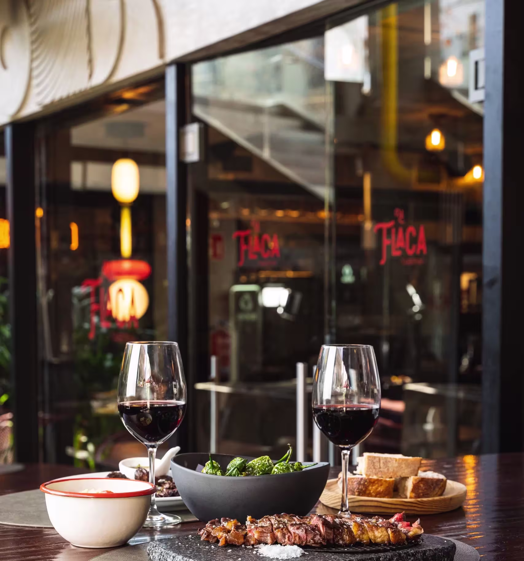 La imagen muestra una mesa de comedor con dos copas de vino tinto, un bol de aceitunas verdes, un pequeño bol con una salsa, rebanadas de pan en una tabla de madera y carne a la parrilla en un plato.