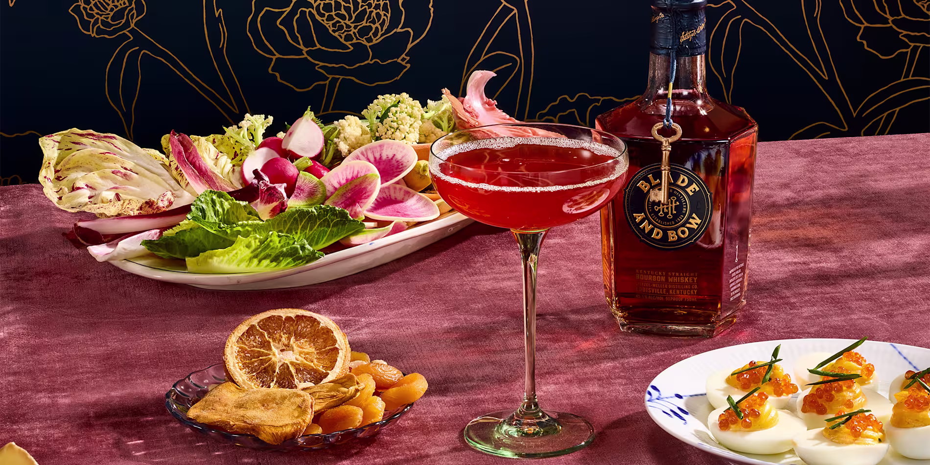 A cocktail in a coupe glass sits beside a bottle of Blade and Bow bourbon, a plate of fresh vegetables, dried fruits, and deviled eggs with garnish on a pink tablecloth.