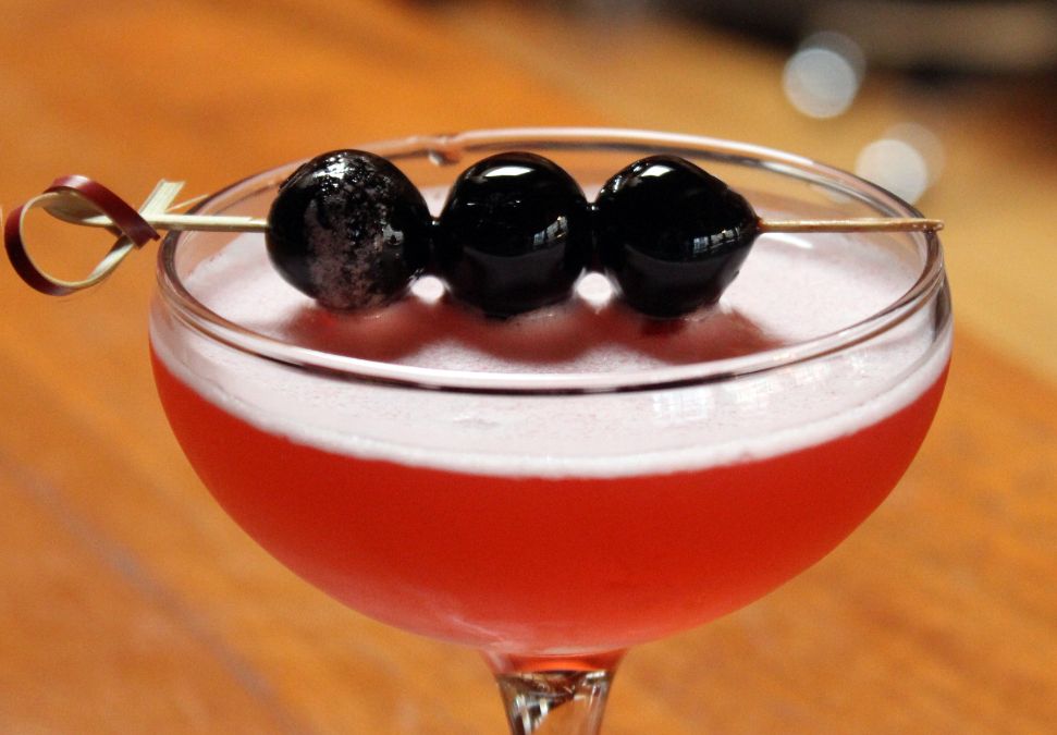 A red cocktail served in a coupe glass, garnished with three dark cherries on a cocktail pick.