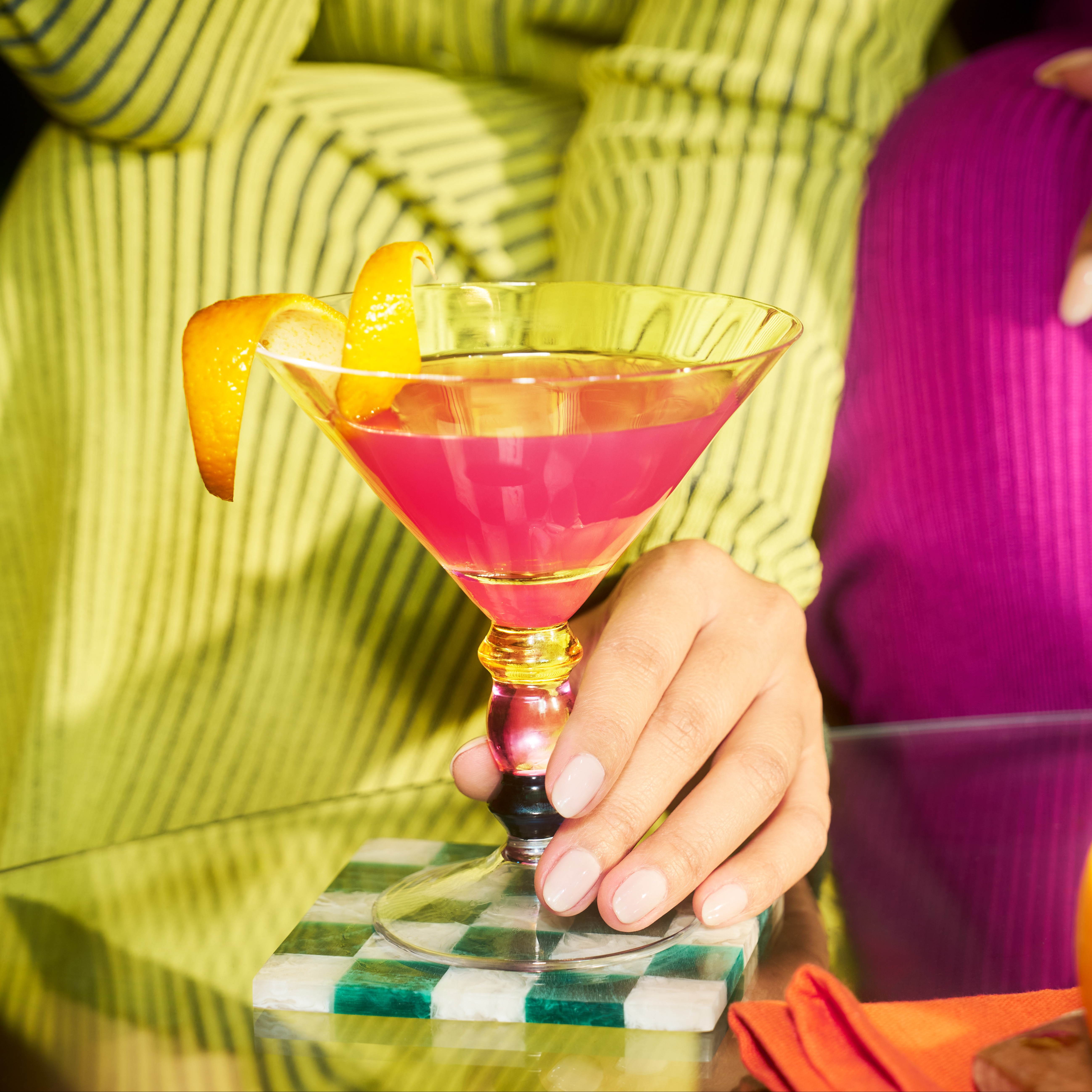 Egy cosmopolitan koktél martinis pohárban, csavart narancshéjjal díszítve, fehér és zöld kockás alátéten egy üvegasztalon.