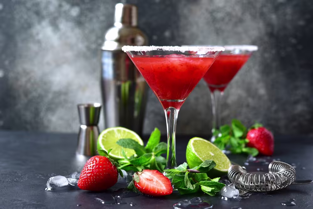 Two strawberry cocktails in martini glasses with sugared rims, surrounded by fresh strawberries, lime, mint, ice cubes, a cocktail shaker, jigger, and strainer on a dark surface.